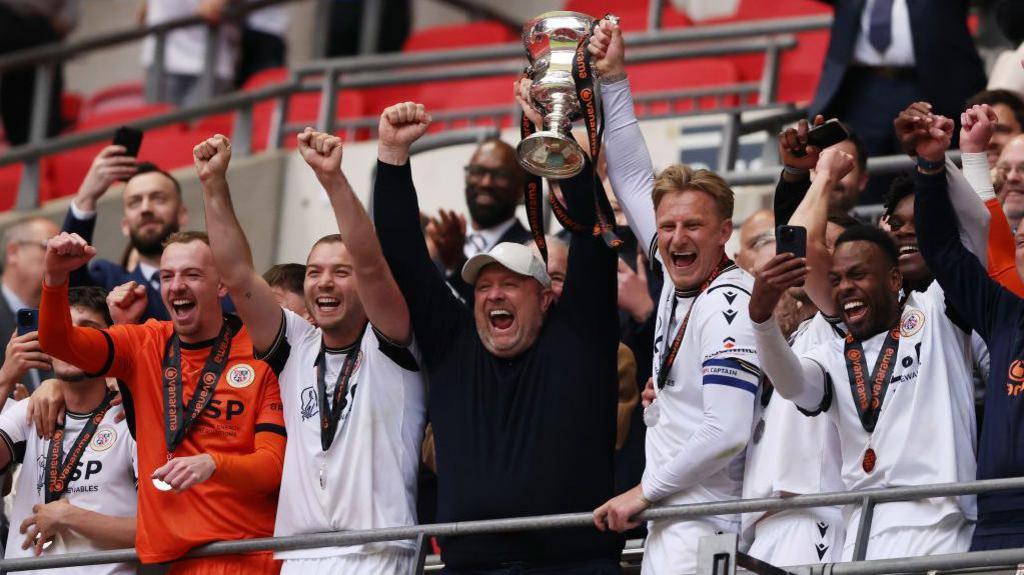 Bromley boss Andy Woodman lifts the National League promotion final winners' trophy, surrounded by Bromley players, in the stand at Wembley Stadium