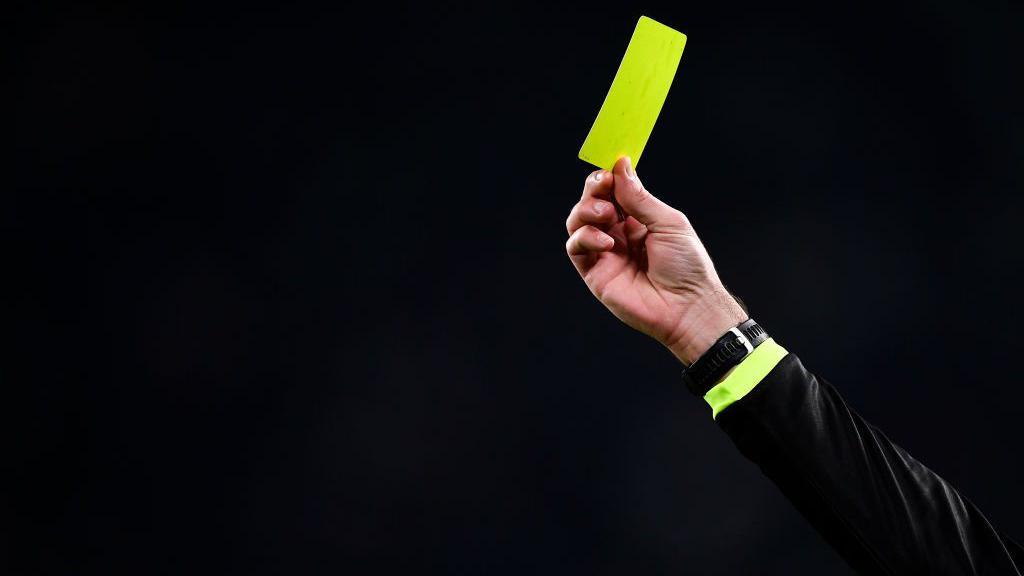 The hand of a football referee is seen showing a yellow card against a black background