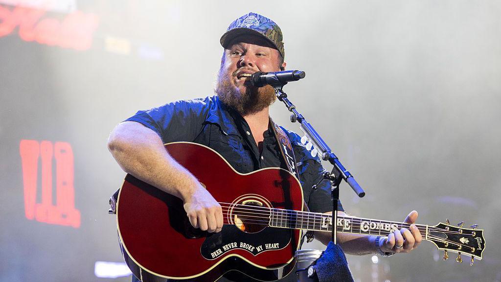 Luke Combs performs during 2025 Austin City Limits Music Festival at Zilker Park. He has a brown beard is wearing a camo baseball cap, wearing a black short sleeve shirt and playing a guitar singing into a microphone.