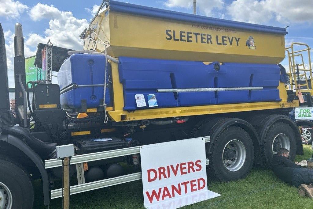 A blue and yellow gritter with the name "Sleeter Levy" on the side. It is pictured at The Driffield Show with a sign saying "Drivers Wanted" placed against it. 