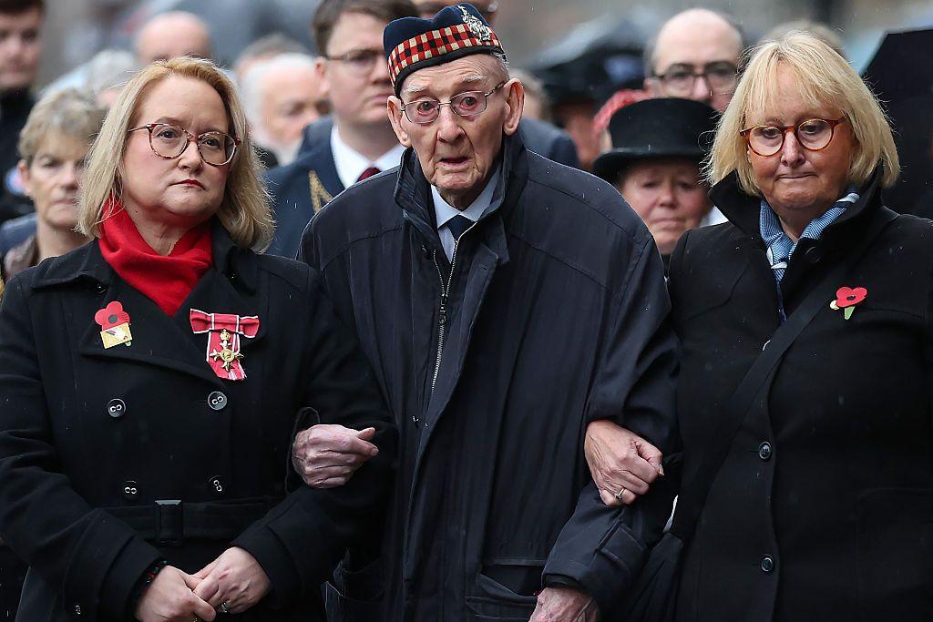 An elderly man wearing a traditional Glengarry bonnet, his arms linked with two women either side of him. The women are both wearing red poppies on their lapels.