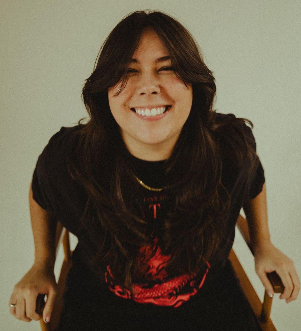 Gigi Perez smiles while looking at camera while sitting in a chair against a neutral background, in a promotional image for her music.