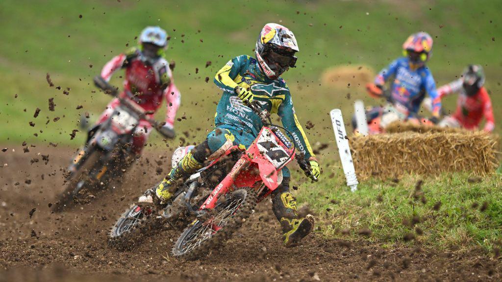 Four people in brightly-coloured protective clothing and helmets, riding motorcycles on a dirt track, with mud flying out behind them.