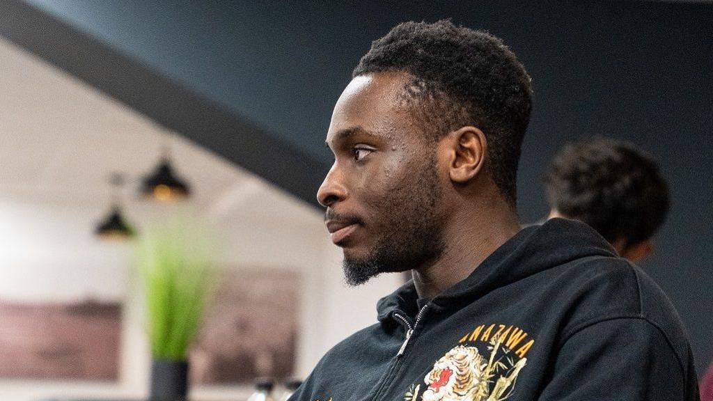 Kamil Conteh pictured looking away from the camera, watching the event. He wears a black hoodie with a large design on one side. 