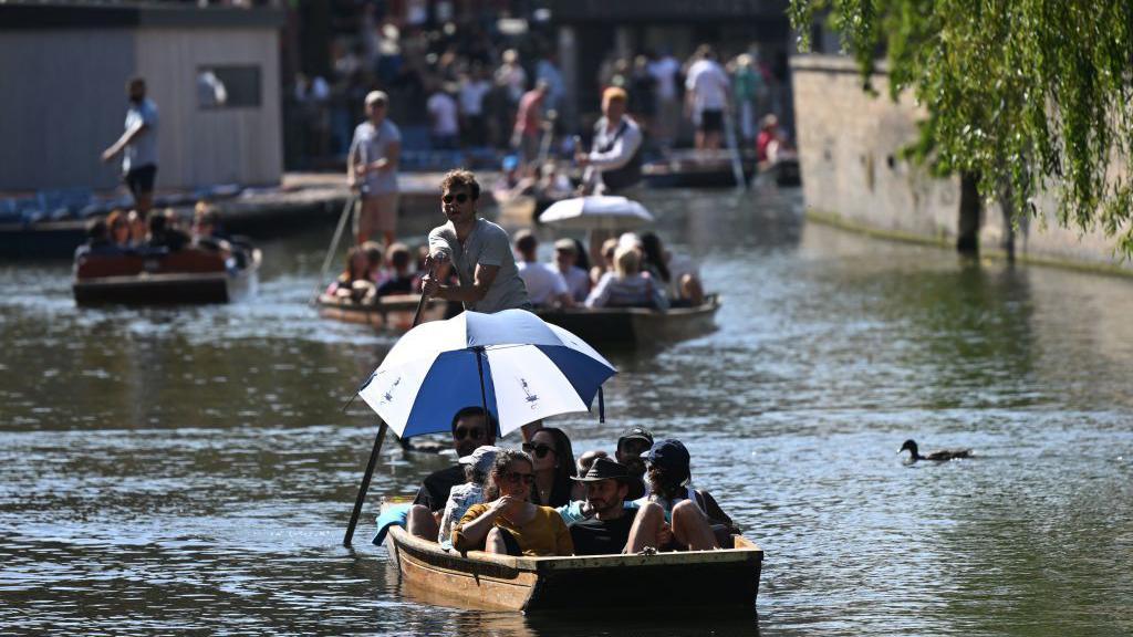 People punting on the River Cam, Cambridgeshire. One person in a punt holds an umbrella to shade themselves from the Sun.