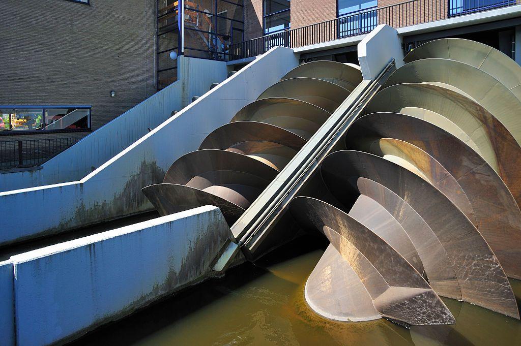 Image of two big metal turbines at a 45 degree angle inside water next to a brick building.