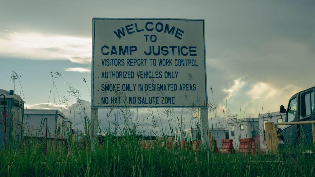 a sign reads "camp justice - visitors report to work control - authorized vehicles only - smoke only in designated areas - no hat - no salute zone" is surrounded by reeds