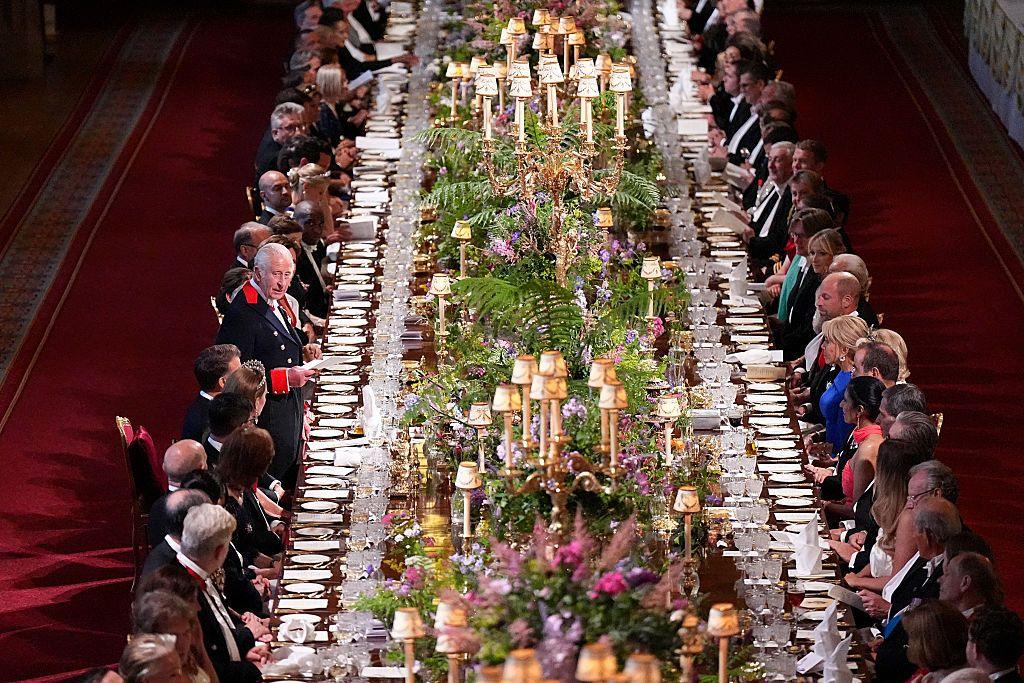 King Charles addresses the banquet's guests, who line a long table covered in flowers, lamps and table-wear.