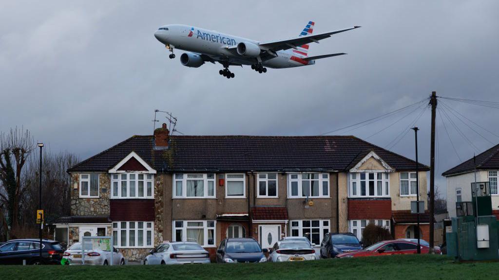 An American Airlines jet flies low over suburban houses, preparing to land under an overcast sky.