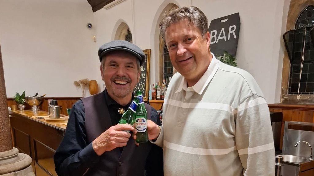 Alan Fletcher and Darren Brooks are in a bar holding bottles of beer smiling to the camera