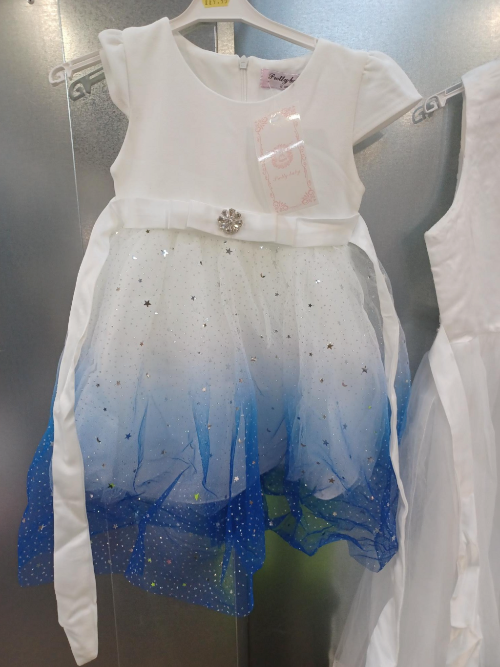 White embellished children's dress with a navy blue gradient trim and chiffon skirt. The dress is hanging on white plastic clothes hanger against a silver metal backdrop. The dress has a white tie-at-the-waist ribbon which is untied and appears to be longer in length than the dress itself.