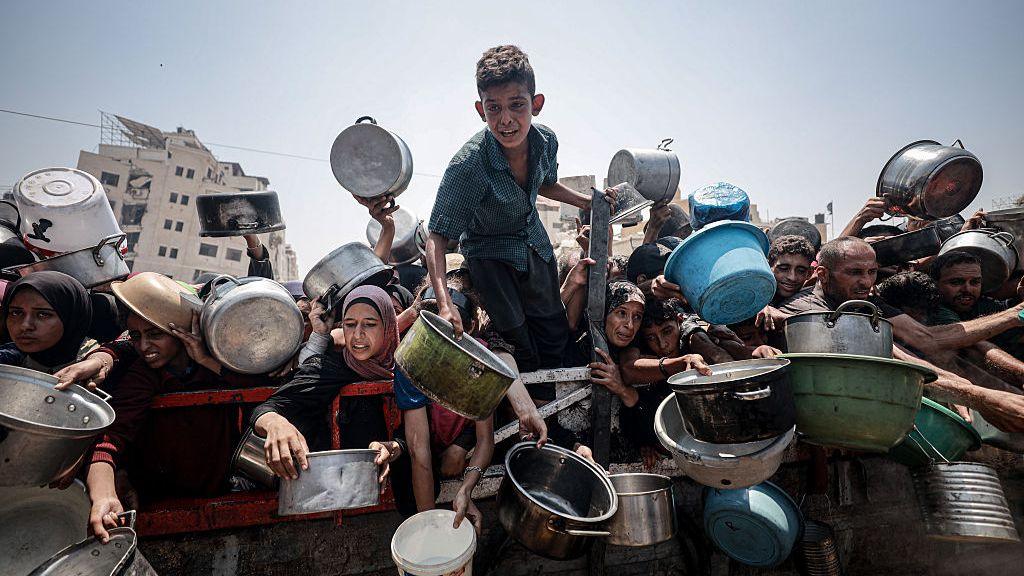 A distressed boy stands above a crowded group of people, many holding bowls and pans, as they attempt to get aid in Gaza. 