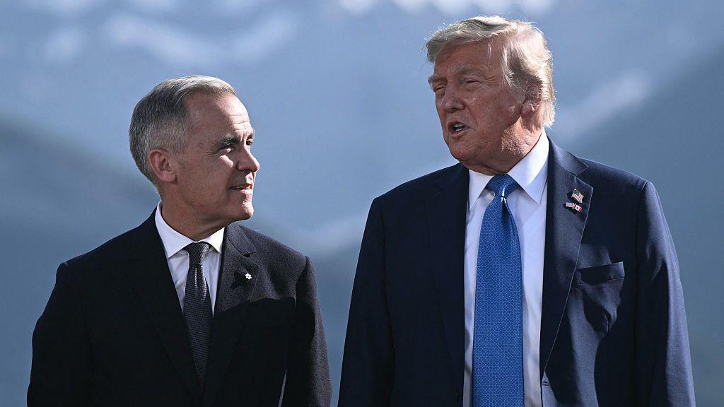 An image of Donald Trump and Mark Carney standing side-by-side at the G7 Summit in Kananaskis, Alberta, in June. The background behind them shows a blurred mountain range. They are both wearing suits, and are facing each other and appear mid-conversation. 
