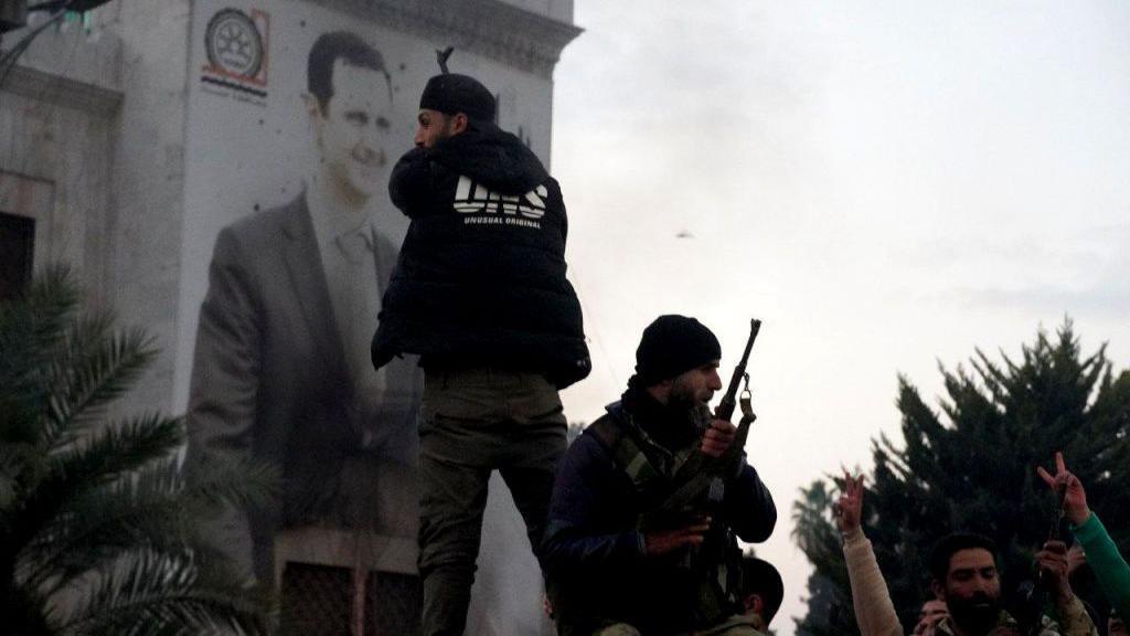 Armed fighters in front of a banner depicting Assad in Hama