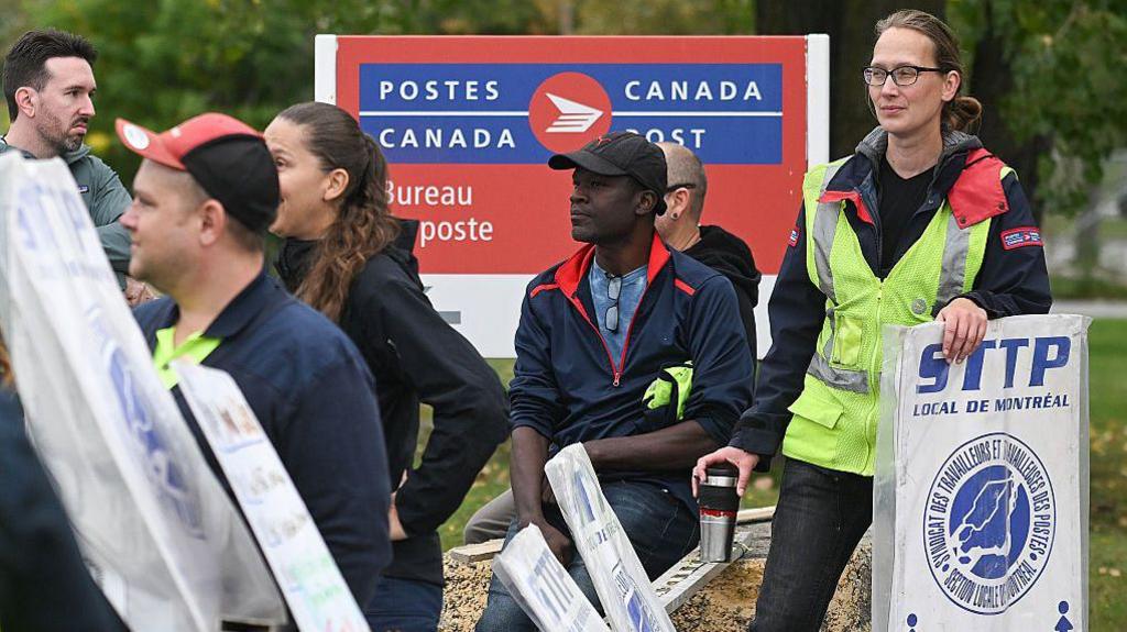 Striking postal workers, some holding white and blue picket signs reading 'STTP, local de Montreal', gather in front of a large blue and red Canada Post post office sign.