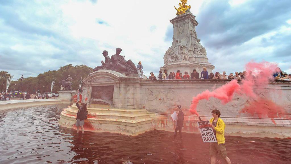 Climate activist from the Animal Rebellion activists group holds a red flare as they walk in the red-colour dyed fountain outside Buckingham Palace 