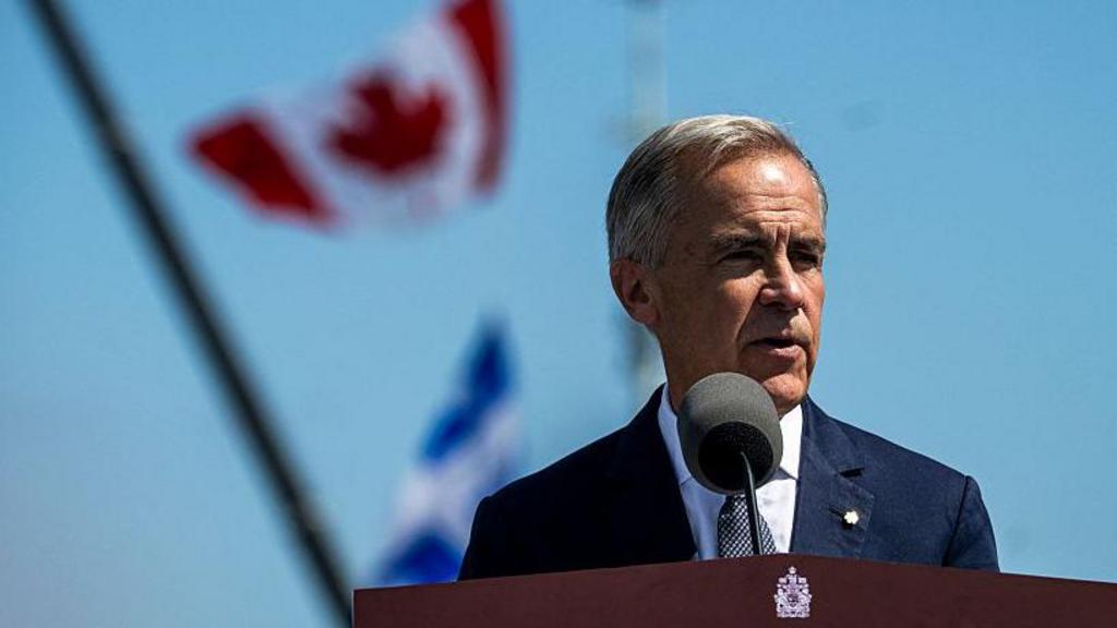 Mark Carney stands at a lectern outside on a sunny day with a Canadian flag behind him