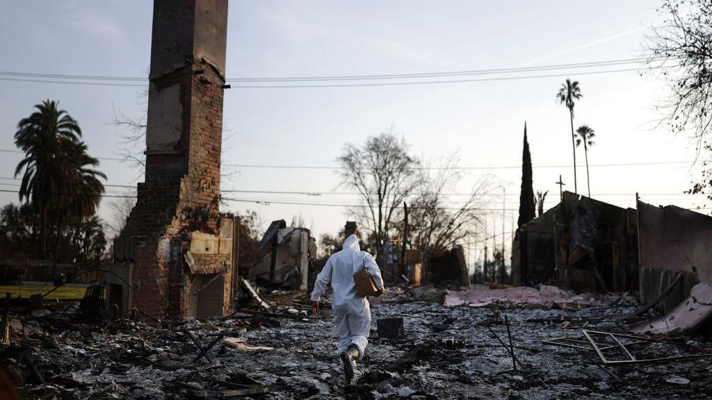 A person in a protective suit walks through the aftermath of the fires in LA. A chimney stands surrounded by debris and charred remains of buildings 