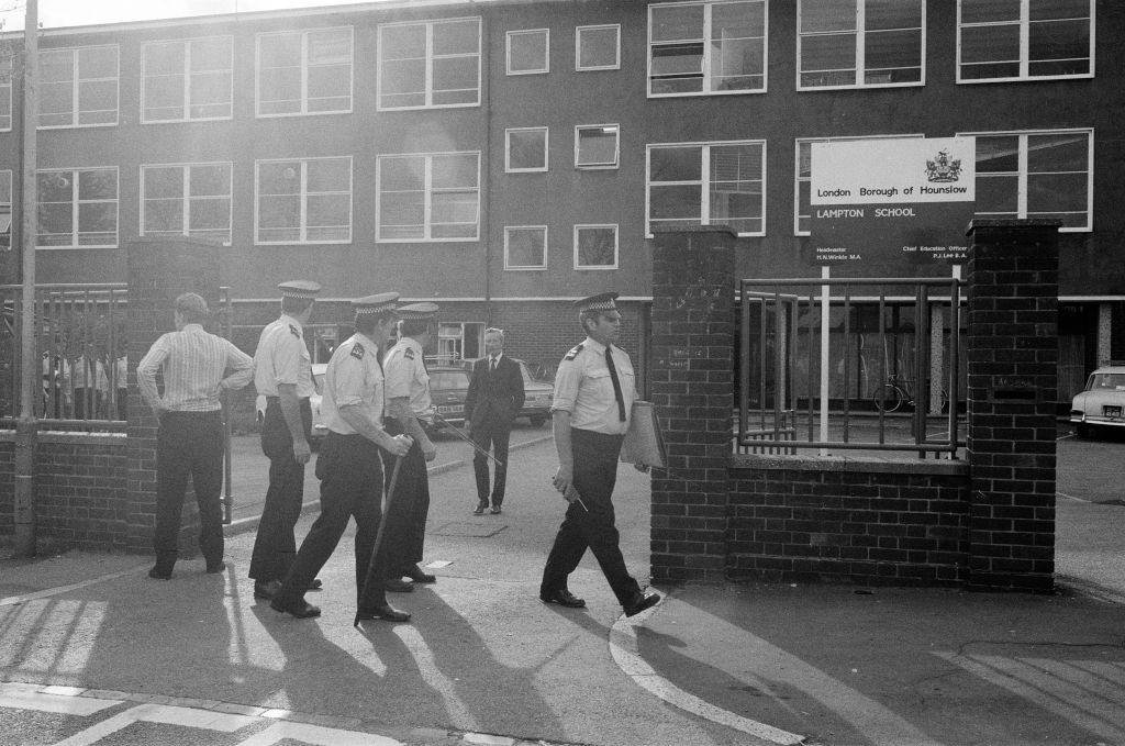 Police officers outside Lampton School. Three uniformed officers stand around holding sticks and another has a walkie-talkie and a file under his name. Two plain clothes officers are also there. The school is a plain, boxy building with lots of square windows, a wall and a fence. The school gates are open.