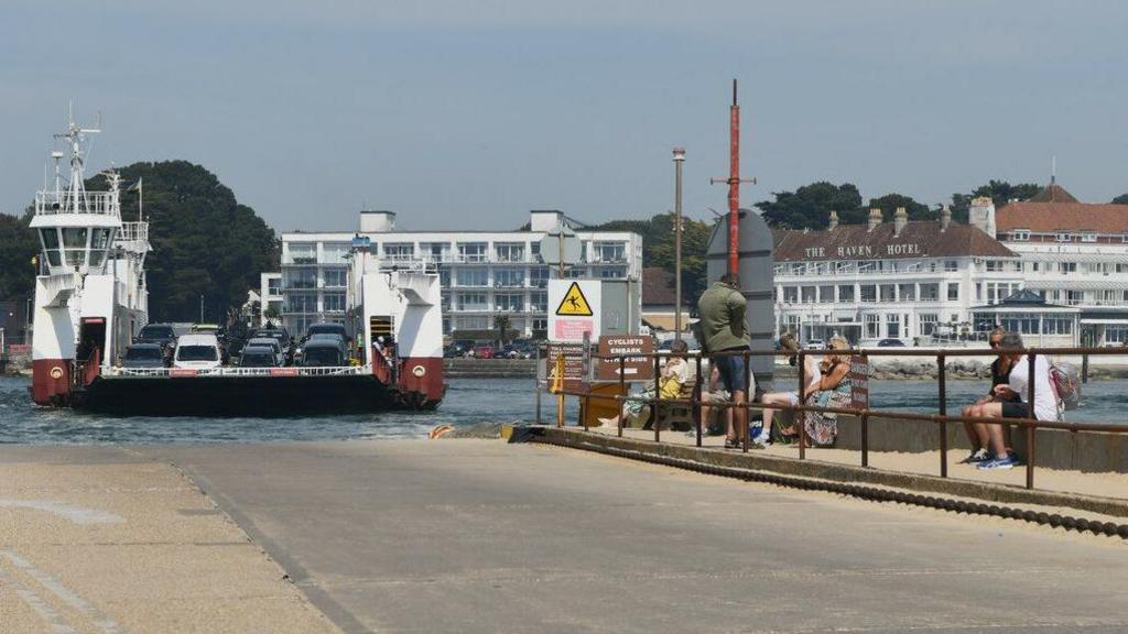 View from the concrete slipway as the Sandbanks Ferry approaches. The vessel is an open-top, roll-on roll-off ferry with high sides and the cabin on the left side. In the background across the water is the white three-storey Haven Hotel.