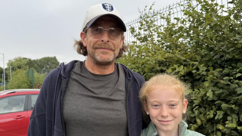 A man and a girl looking at the camera. The man is wearing a white baseball cap and a grey T-shirt. The girl is wearing a green fleece jacket. They are standing in front of a hedge, a fence and a red car.