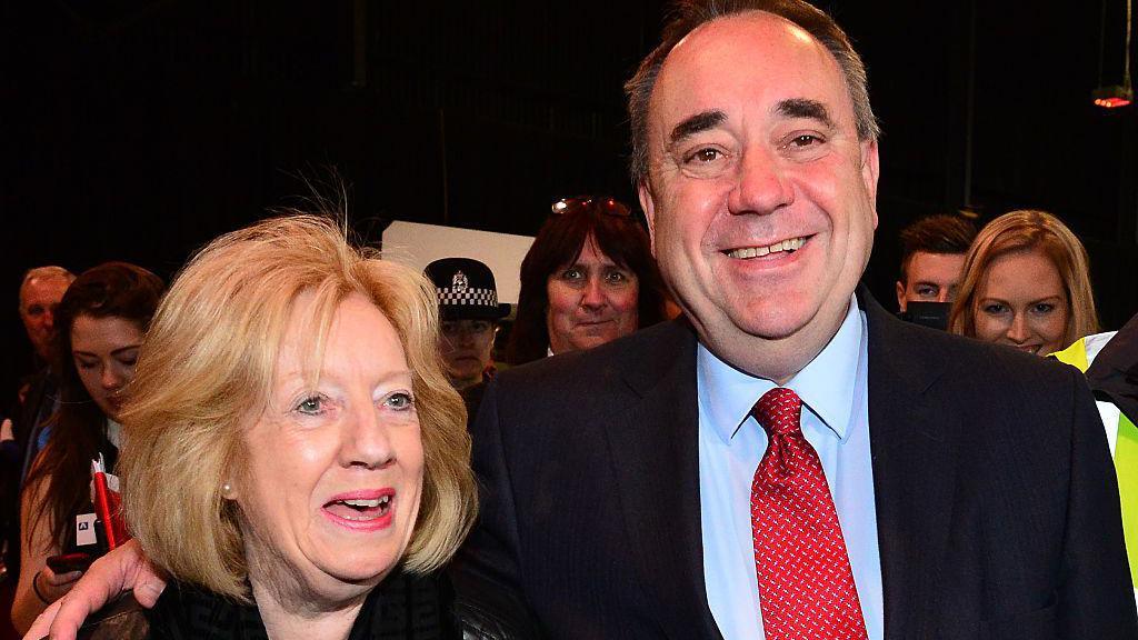 Alex Salmond pictured with his wife Moira during the 2015 UK Parliamentary elections. They are smiling and he has his arm around her. He is wearing a blue shirt and red tie with a dark blue suit.