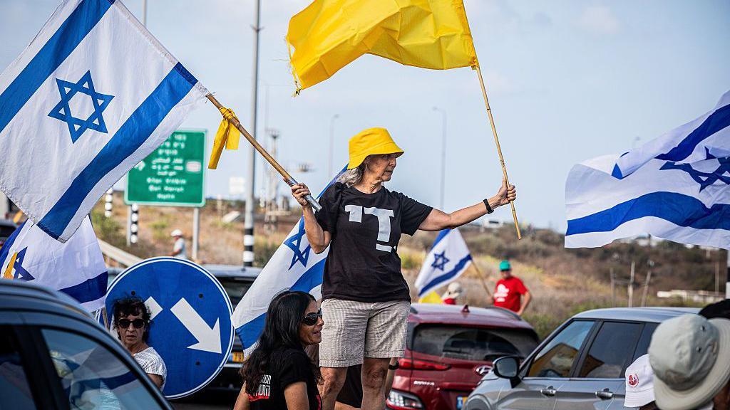 A woman holding an Israeli flag and a yellow flag representing the hostages waves them while stood in the middle of a busy road at a daytime protest. 