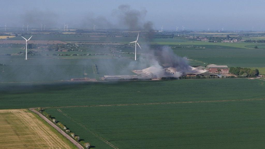 Smoke billowing from a fire at a farm building. There are crop fields in the foreground and wind turbines visible behind the farm.