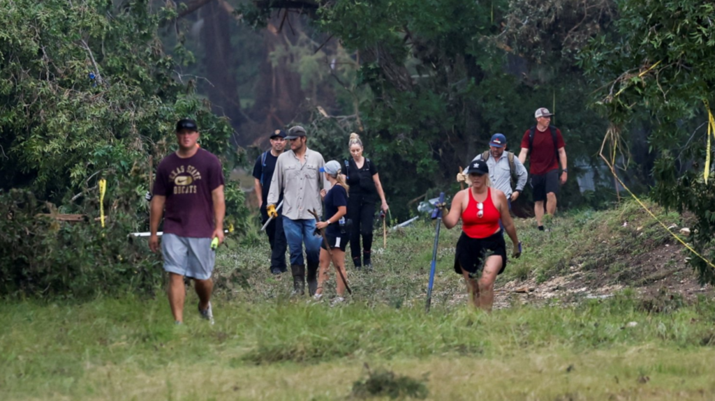 Five men in and three women search along a muddy grass path holding sticks 