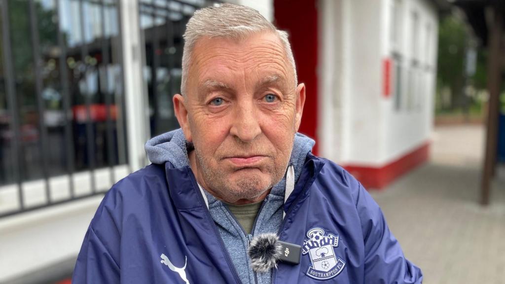 A Saints fan wearing a Southampton FC jacket and pin badge looks upset. He is sitting outside a put. 