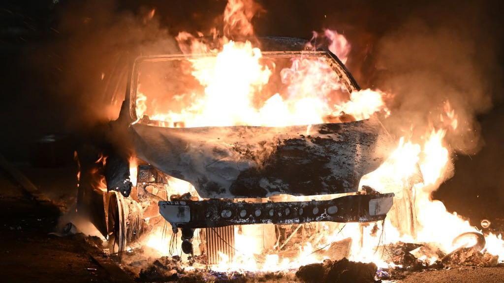 Car on fire during the Ely riots, with flames surrounding it