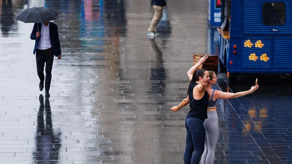 wet street with a man walking along with an umbrella and two women posing for selfie in the rain