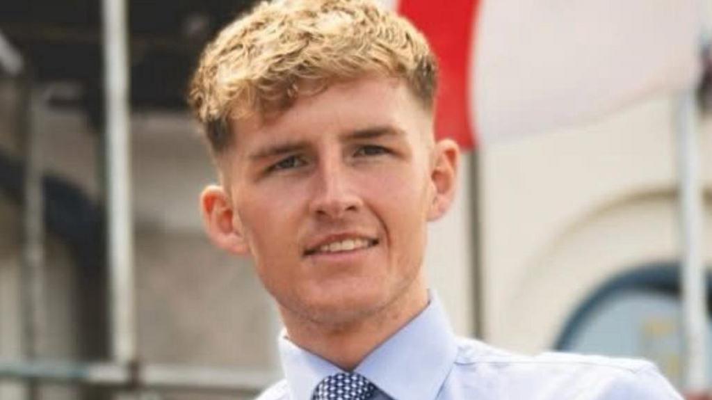 Oliver Freeston is smiling into the camera. He has blonde curly hair and is wearing a white shirt with a blue and white tie. He's stood in front of a building with a flag flying behind him.