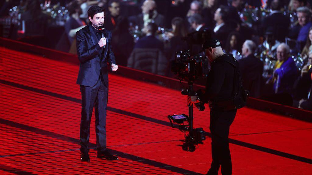 Actor and comedian Jack Whitehall speaking into an F1-branded microphone with a cameraman filming him 