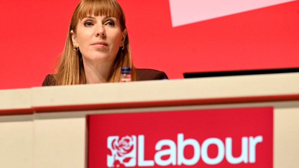 Labour Deputy Prime Minister Angela Rayner at the Labour Party Conference. She is sitting at a long table behind a red Labour logo, and in front of a red backdrop