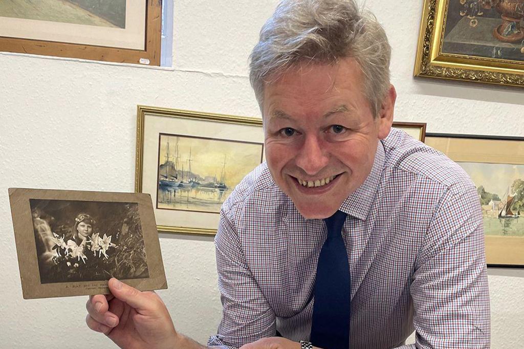 A man with grey hair, a purple and white square patterned shirt and navy tie is holding a black and white photo of a girl with fairies in front of her. Behind him are paintings on a wall.