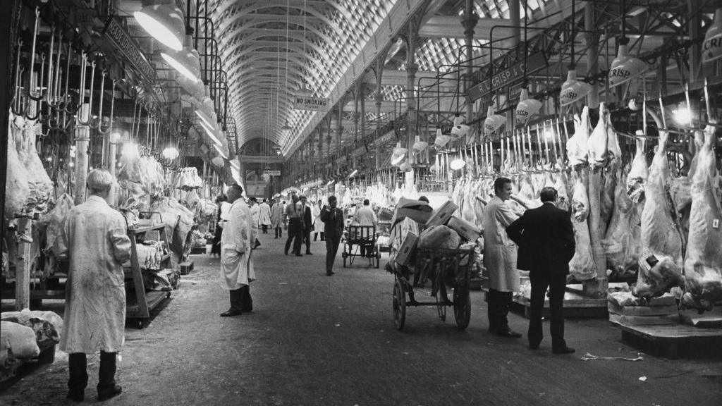 Rows of animal carcasses hang from hooks, beside market traders, porters and shoppers at Smithfield Meat Market circa 1965 