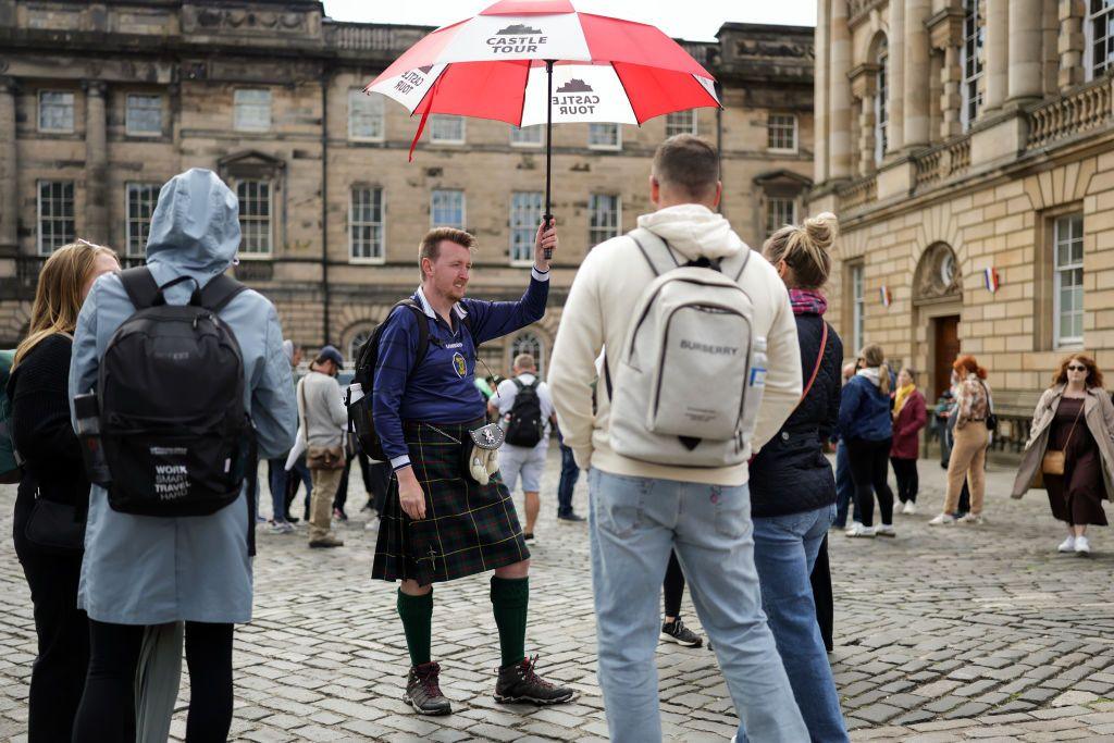 A kilted man with an umbrella speaks to tourists in a cobbled street