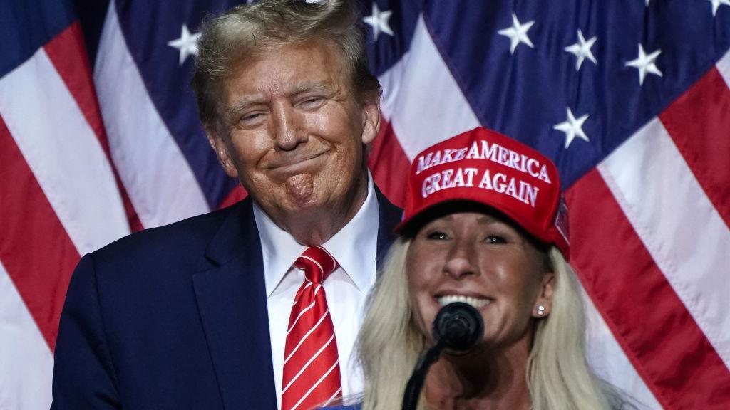 Congresswoman Marjorie Taylor Greene, wearing a red cap and speaking into a microphone speaks in support of Trump at his campaign rally in Rome, Georgia in March 2024. Trump looks on in the background, smiling.