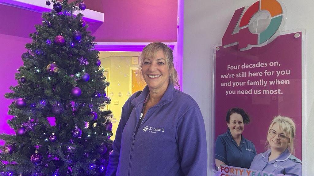 Teresa Bickle wearing a blue fleece jacket with the St Luke's logo on the right. She is smiling towards the camera and there is a Christmas tree on the left.