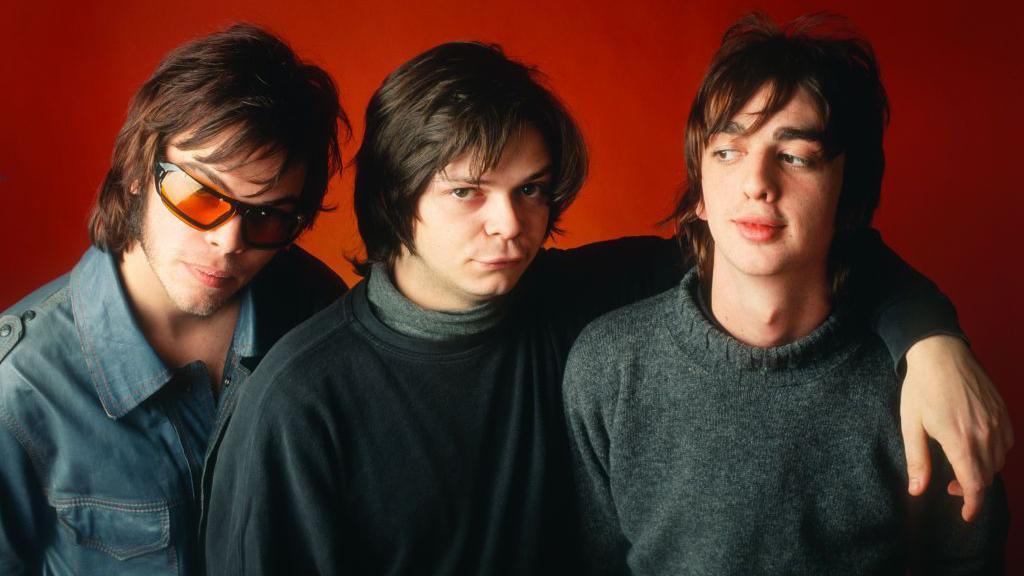 Gaz Coombes, wearing orange shades and a denim jacket, Mick Quinn, wearing a black jumper and a polar necked shirt, and Danny Goffey, wearing a grey woolly jumper, standing in front of an orange background