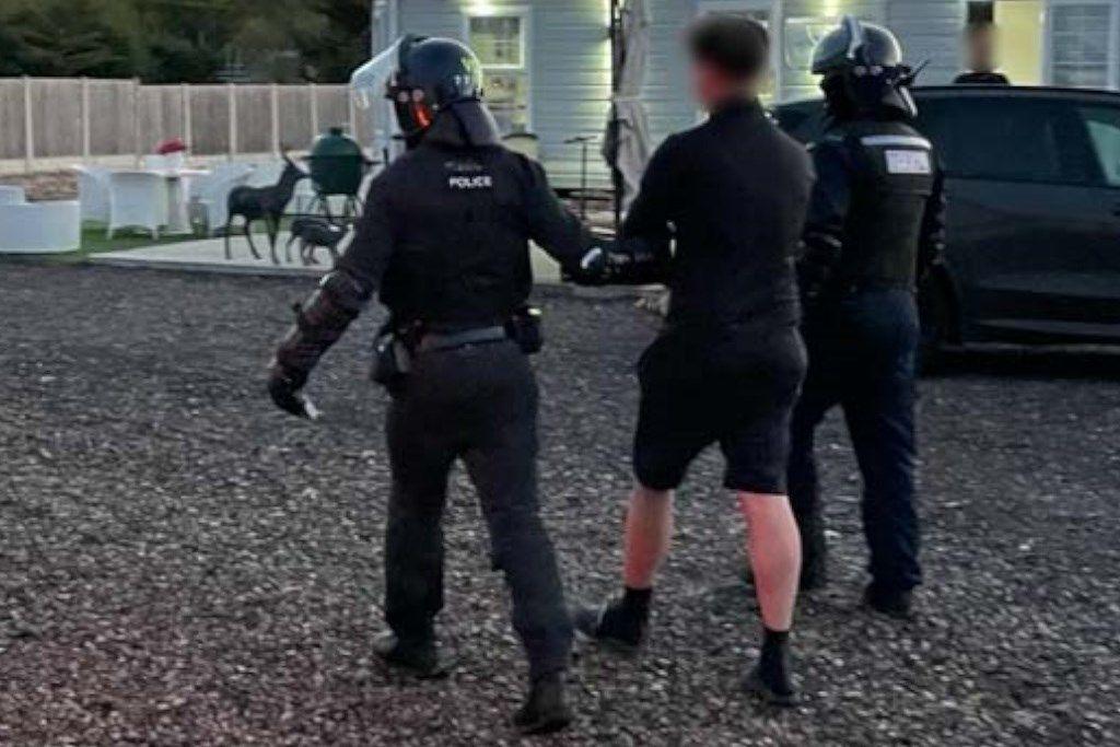 Shot from behind of one of the gang members being led away, with a police officer at either side. The back of his head is blurred out and he is wearing a black fleece, shorts, ankle socks and slider. The officers wear protective raid uniform. They walk along a pebbled path with a caravan and car partially seen in the background.