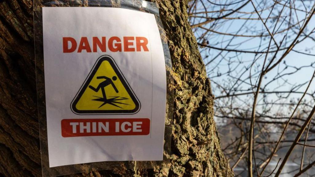 A white paper sign on a tree reads "Danger Thin Ice" with a hazard graphic of a person falling. Branches and a body of water can be seen in the background. 