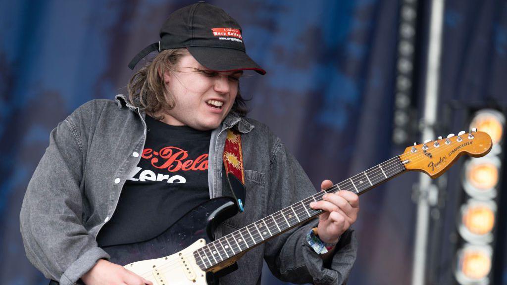 Zac Schulze is wearing a black T-shirt, with a grey overshirt and a black cap. He is playing a guitar 