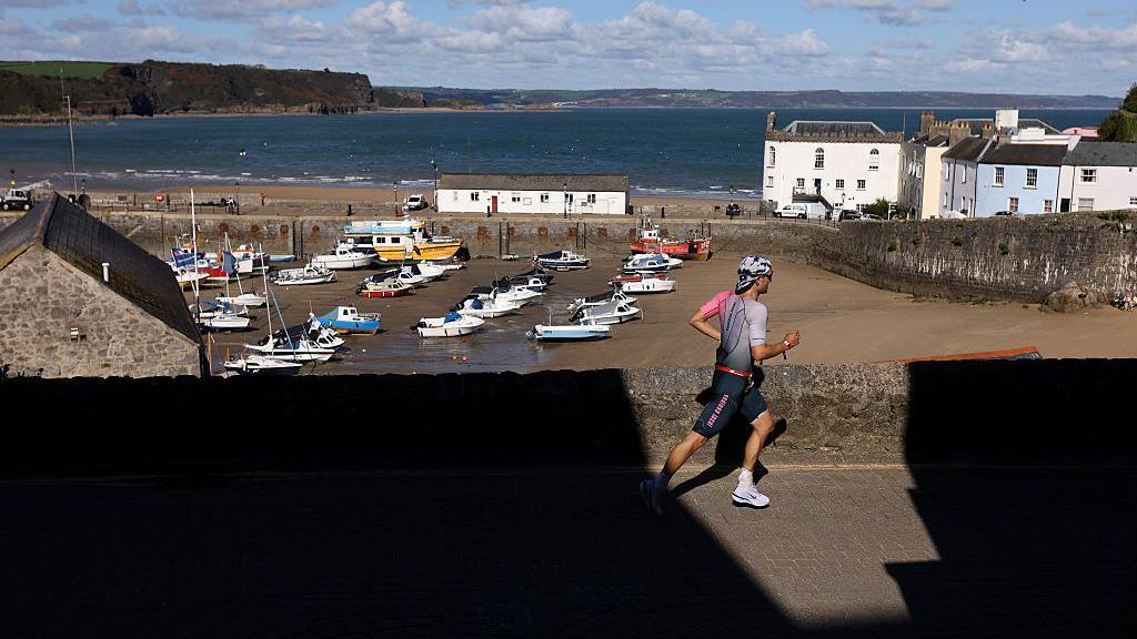 A man runs alongside a wall overlooking Tenby harbour with white houses and boats on the sand. The sea is behind.