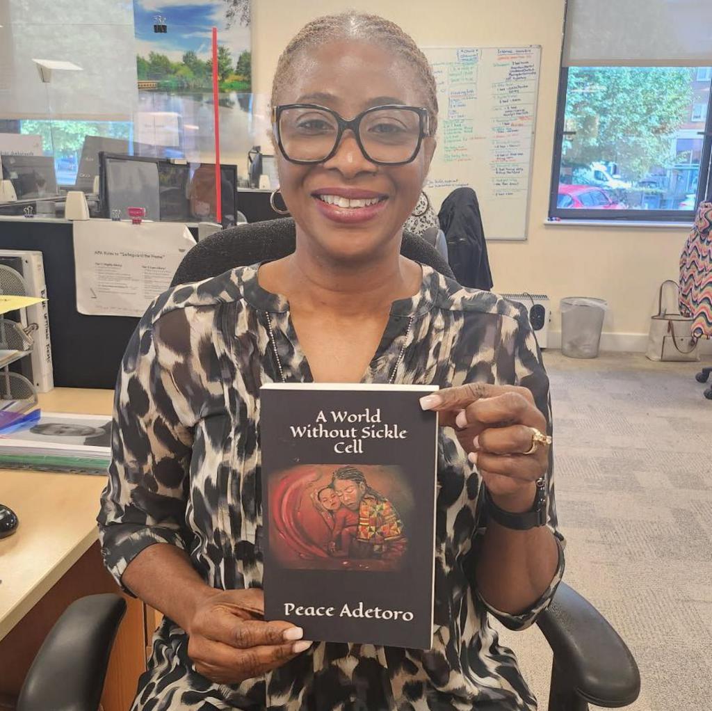 A woman is holding a book to the camera which has the title "A world without sickle cell" and the author's name: Peace Adetoro