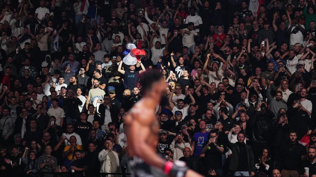 Morgan Charriere celebrates on the cage with a spotlight on a fan holding a French flag