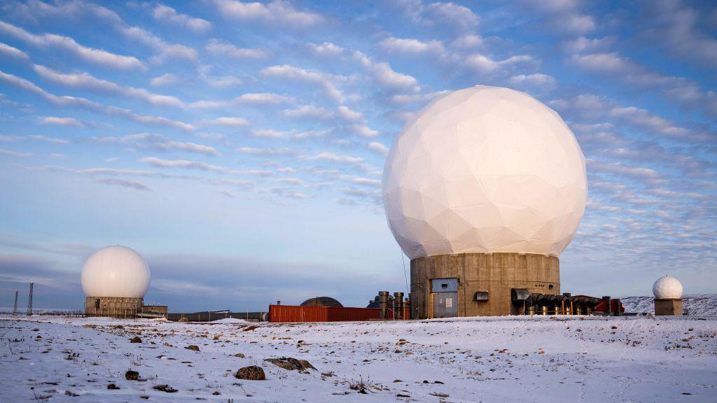 A remote Arctic landscape in northern Greenland, featuring the Pituffik Space Base (formerly Thule Air Base). The image shows three massive white geodesic radar domes positioned on a snow-covered plain. The largest dome is centrally located on a concrete structure, surrounded by other domes.