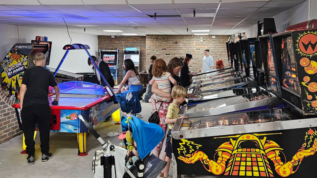Inside the arcade with several brightly coloured pinball machines lined up against one wall and people, including children, playing on them. There is also an air hockey table with young people playing.