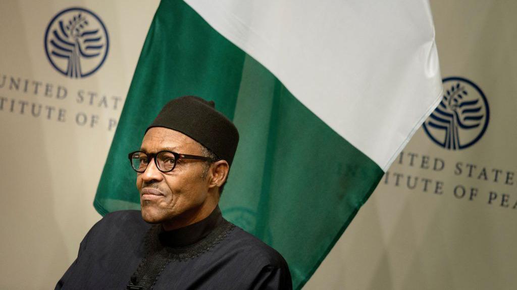 Muhammadu Buhari sits in front of a Nigerian flag and signs that say United States Institute of Peace. He wears a black top, black cap and glasses.